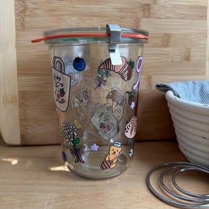 May include: A clear glass Weck jar with a glass lid, orange rubber seal, and metal clamp. The exterior features colorful stickers of a bear with honey, flowers, a croissant, a blueberry, and a shopping bag with the text shop small.