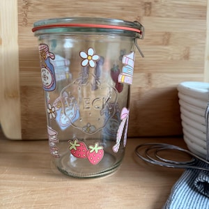 May include: A clear glass Weck jar with a red rubber seal and metal clamps. The jar is decorated with colorful stickers of strawberries, flowers, books, and bows. The jar is sitting on a wooden surface.