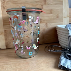 May include: A clear glass jar with a red rubber seal and metal clamp closure. The jar is decorated with colorful illustrations of cowboy boots, butterflies, and flowers. The word "WECK" is embossed on the front.