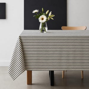 May include: A rectangular tablecloth featuring a pattern of thin, horizontal dark grey stripes on a light cream background. A clear glass vase containing white gerbera daisies and green stems sits on the surface of the covered table.