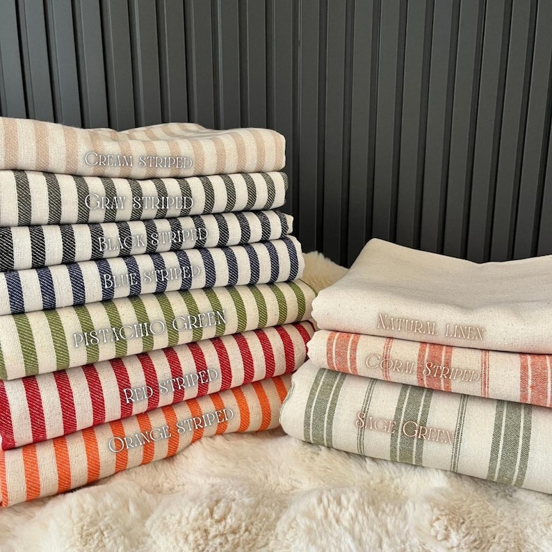 May include: A stack of striped fabric swatches in various colors, including cream, gray, black, blue, green, red, and orange. The swatches are labeled with their corresponding colors.  The fabric is a natural linen blend.