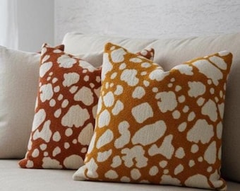 Puede incluir: Dos cojines decorativos cuadrados con un patrón blanco y naranja. Los cojines están colocados sobre un sofá beige. El patrón se asemeja a un diseño moteado, agregando interés visual a la habitación. Un toque cálido.