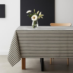 May include: A striped tablecloth in black and beige covers a table. A glass vase with white flowers is on the table. The tablecloth's pattern is visible on the table's edge. The table is made of wood.