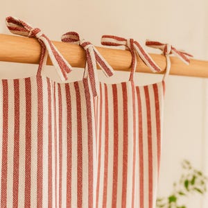 May include: Red and white striped curtains with matching tie closures. The curtains are hanging from a wooden rod, creating a classic and cozy aesthetic. The fabric has a textured appearance.