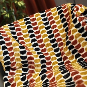 May include: Close-up of a fabric featuring a repeating pattern of semi-circles in black, gold, rust, and cream. The textured fabric is draped, creating folds and shadows that enhance the design's depth and visual interest.