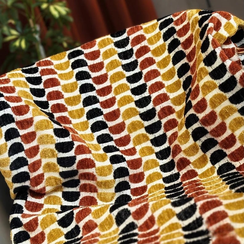 May include: Close-up of a fabric featuring a repeating pattern of semi-circles in black, gold, rust, and cream. The textured fabric is draped, creating folds and shadows that enhance the design's depth and visual interest.