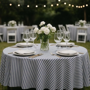 Op de afbeelding: Een ronde tafel gedekt voor een buitenevenement, bedekt met een blauw-wit gestreept tafelkleed. De tafel is gedekt met borden, servetten, goudkleurig bestek en wijnglazen. Een vaas met witte bloemen staat in het midden.