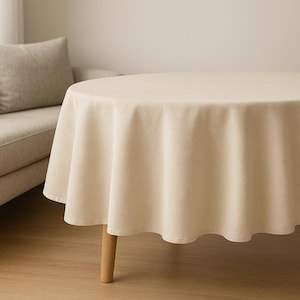 May include: A round, cream-coloured tablecloth on a wooden-legged table. The tablecloth has a textured finish and hangs with soft folds. The table is in a room with a neutral colour scheme.
