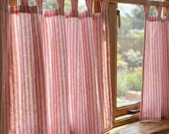 May include: Pink and white striped curtains hanging in front of a window. The curtains are made of a lightweight fabric and are attached to a wooden rod with fabric loops. The window frame is made of wood, and the background shows a view of the outdoors.