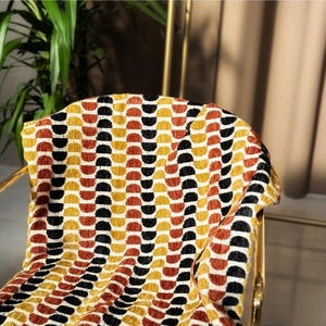 May include: A gold-coloured chair with a patterned throw. The throw features a repeating design of semi-circles in shades of yellow, orange, black, and white. The chair's frame is visible, adding a touch of elegance to the scene.