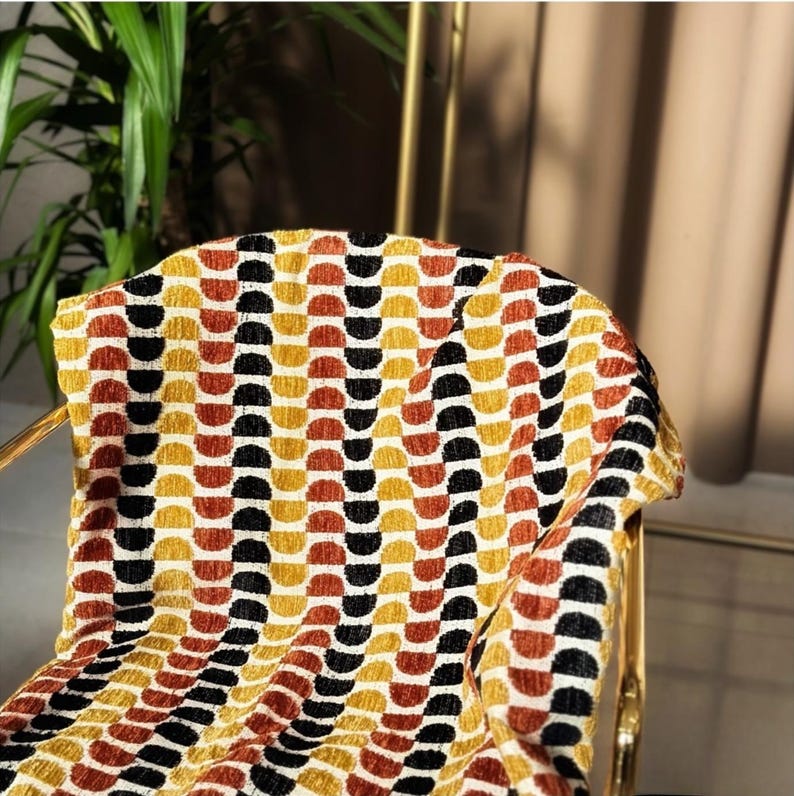 May include: A gold-coloured chair with a patterned throw. The throw features a repeating design of semi-circles in shades of yellow, orange, black, and white. The chair's frame is visible, adding a touch of elegance to the scene.