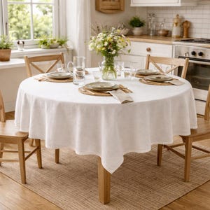 Könnte beinhalten: Ein runder Esstisch für vier Personen mit weißer Tischdecke, Holzstühlen und gewebten Tischsets. Eine Vase mit Blumen steht in der Mitte. Der Tisch steht auf einem gewebten Teppich.