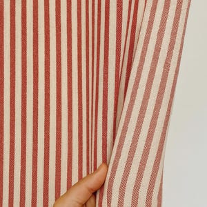 May include: Close-up of a fabric with vertical stripes in red and off-white. The fabric is being held, revealing the texture and pattern. The stripes are evenly spaced, creating a classic design.
