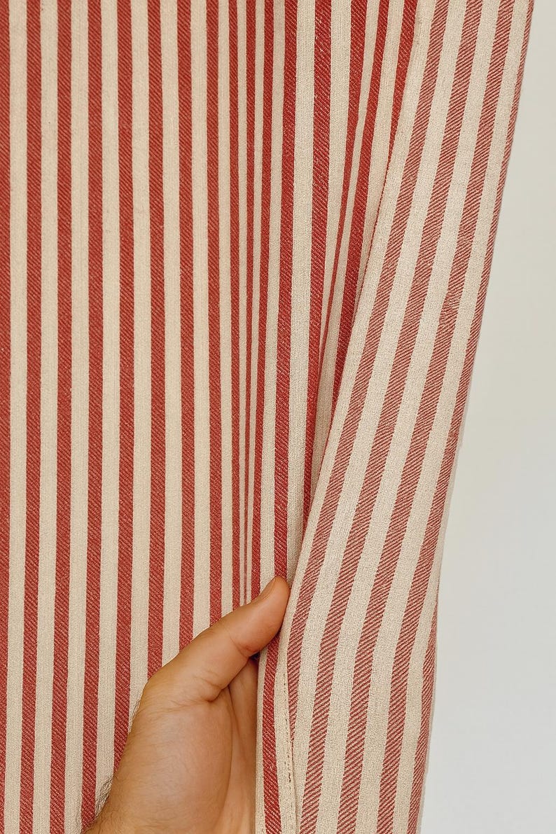 May include: Close-up of a fabric with vertical stripes in red and off-white. The fabric is being held, revealing the texture and pattern. The stripes are evenly spaced, creating a classic design.