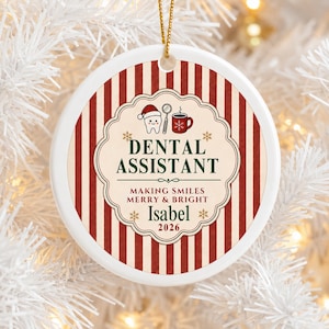 May include: A round white ceramic ornament featuring a red and white vertical striped background. The centre displays a cream-coloured scalloped label with a tooth wearing a Santa hat, a dental mirror, a mug, and the text DENTAL ASSISTANT, MAKING SMILES MERRY & BRIGHT, Isabel, and 2026.