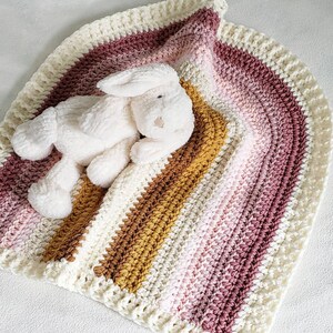 May include: A crocheted baby blanket with a pink, white, and brown ombre design. A white plush bunny is lying on top of the blanket.