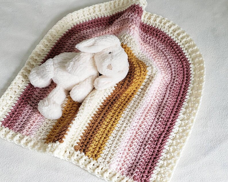 May include: A crocheted baby blanket with a pink, white, and brown ombre design. A white plush bunny is lying on top of the blanket.