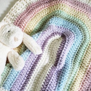 May include: A white crocheted rainbow blanket with a plush white bunny toy on top. The blanket is made with a soft, textured yarn and features a rainbow of colors, including pink, yellow, green, blue, and purple.