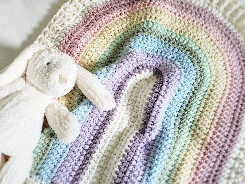 May include: A white crocheted rainbow blanket with a plush white bunny toy on top. The blanket is made with a soft, textured yarn and features a rainbow of colors, including pink, yellow, green, blue, and purple.
