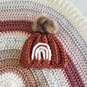 May include: A crocheted baby hat with a brown and white rainbow design and a brown faux fur pom pom.