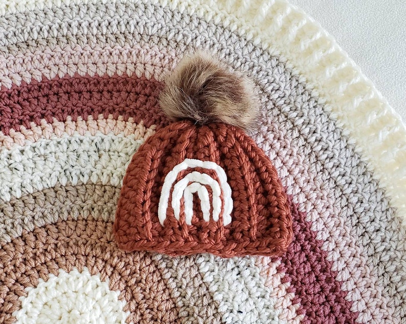 May include: A crocheted baby hat with a brown and white rainbow design and a brown faux fur pom pom.