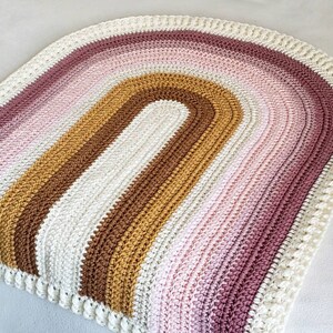 May include: A crocheted rainbow rug with a white border. The rug is made with a variety of colors, including pink, brown, and gold.