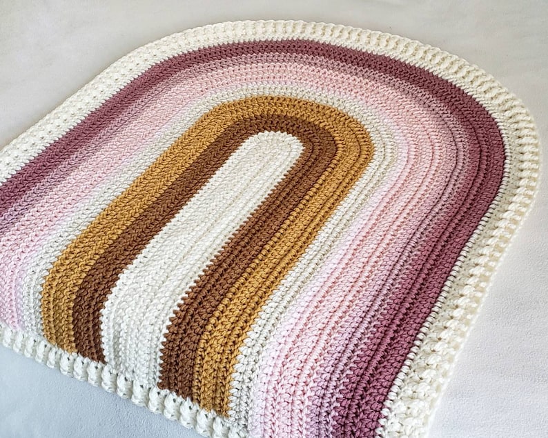 May include: A crocheted rainbow rug with a white border. The rug is made with a variety of colors, including pink, brown, and gold.