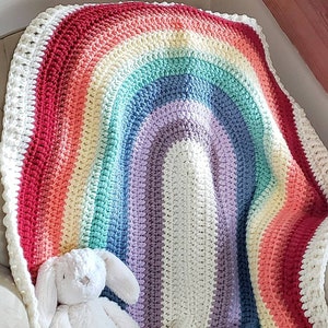 May include: A crocheted rainbow blanket with a white border. The blanket is folded in half and draped over a chair.