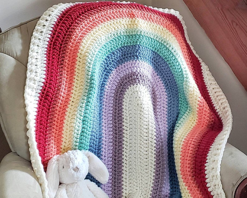 May include: A crocheted rainbow blanket with a white border. The blanket is folded in half and draped over a chair.
