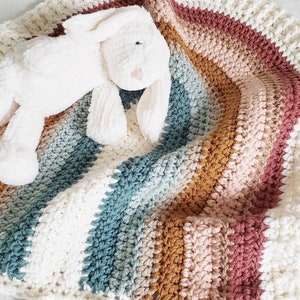 May include: A white crocheted baby blanket with stripes of pink, brown, and blue. A white plush bunny is lying on top of the blanket.