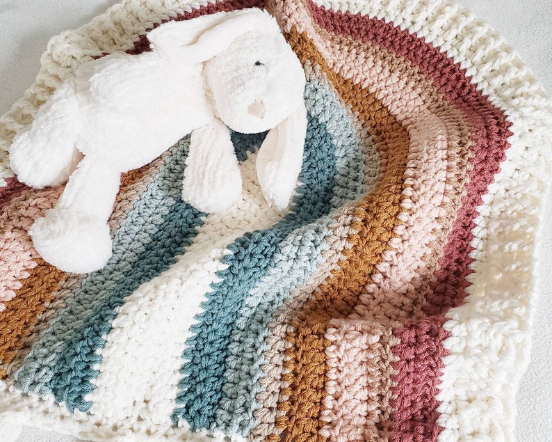 May include: A white crocheted baby blanket with stripes of pink, brown, and blue. A white plush bunny is lying on top of the blanket.