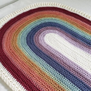 May include: A crocheted rainbow rug in shades of red, orange, yellow, green, blue, purple, and white. The rug is oval shaped and has a white border.