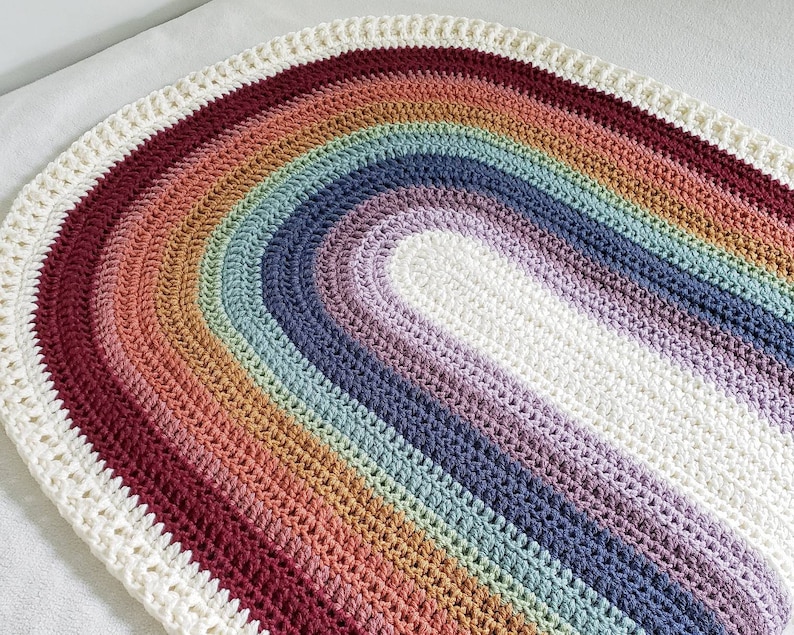 May include: A crocheted rainbow rug in shades of red, orange, yellow, green, blue, purple, and white. The rug is oval shaped and has a white border.