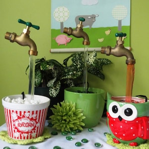 May include: Three decorative tabletop fountains with water flowing from brass taps into a popcorn bucket, a green planter, and an owl-shaped planter. The fountains are on a white surface with green glass stones scattered around.