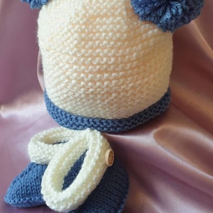 May include: A knitted baby hat and booties set in white and blue. The hat has two blue pom-poms on top. The booties have a white cuff and a blue sole.