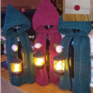 May include: Three wine bottles dressed in knitted hooded capes, two in dark green and one in burgundy. Each cape features a small, light-up lantern hanging from a cord. An inset shows a cream-colored envelope sealed with red wax.
