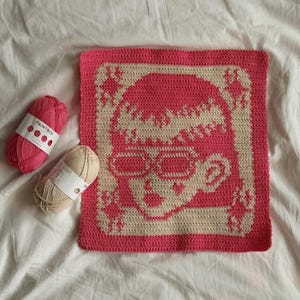 Puede incluir: Un cuadrado de ganchillo rosa y crema con un diseño de calavera con gafas. La calavera está rodeada por un borde crema con detalles de estrella. Dos ovillos de lana, uno rosa y otro crema, están junto al cuadrado.