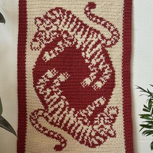 May include: A crocheted wall hanging featuring a red and white tiger design. The tiger is depicted in a curled position with its tail wrapped around its body. The wall hanging is framed with a red border and has a wooden dowel at the top.