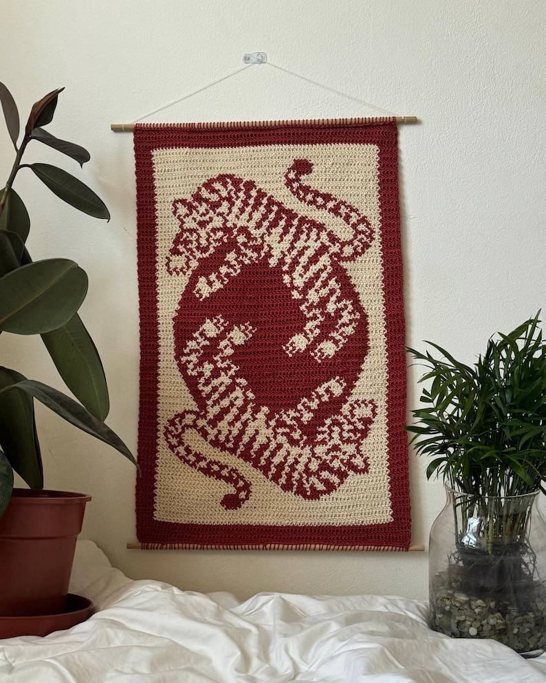 May include: A crocheted wall hanging featuring a red and white tiger design. The hanging is framed with a wooden dowel rod and white string.