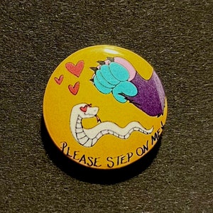 Op de afbeelding: Een gele knop met een witte slang met rode harten als ogen, een blauwe pootafdruk en de tekst "PLEASE STEP ON ME".