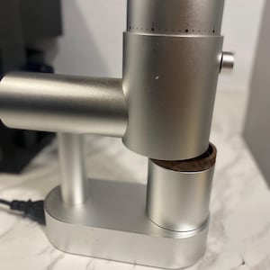May include: A silver electric coffee grinder with a cylindrical body, a horizontal motor housing, and a vertical adjustment dial featuring numerical markings. The device includes a small, dark wood-rimmed collection cup positioned beneath the main unit.