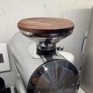 Puede incluir: Un molinillo de café con cuerpo de acero inoxidable cepillado y tapa circular de madera oscura. La parte superior es un embudo de cromo pulido sobre la carcasa cilíndrica principal. Se observa una pequeña perilla de ajuste plateada en el lateral.