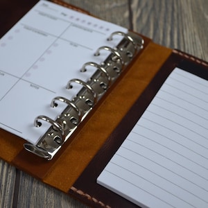 May include: Open leather planner with a silver binder mechanism. The planner is brown with a lighter brown interior and contains white lined paper and a weekly calendar. The planner is open on a wooden surface.