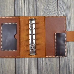 May include: Open leather binder in shades of brown. The binder has a card slot and a metal ring mechanism. The leather is stitched with a contrasting thread. A snap closure secures the binder.