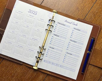 Puede incluir: Agenda de cuero abierta con un calendario de 2025 y páginas de objetivos anuales. El calendario muestra los meses con texto azul. Un bolígrafo azul descansa en el lado derecho. La agenda está abierta sobre una superficie de madera.