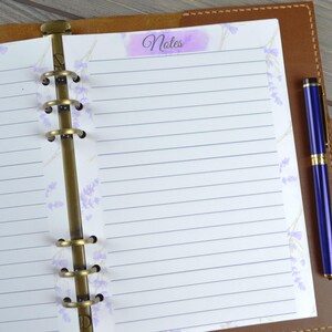 May include: A brown leather bound planner with a brass ring binder. The planner has a white page with lined paper and a lavender floral design. The page has a purple tab with the word "Notes" written on it. A blue pen is placed on the right side of the planner.