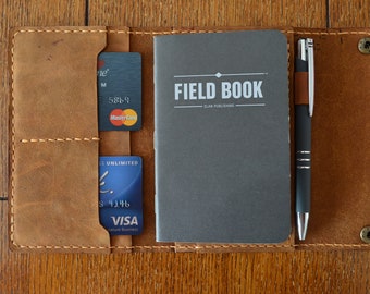 May include: A brown leather notebook cover with a pen holder, two card slots, and a black notebook with the text "FIELD BOOK" and "ELAN PUBLISHING".