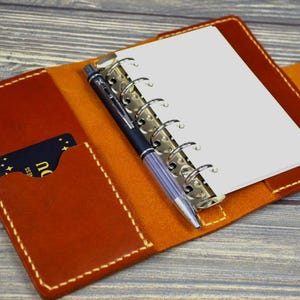 May include: A brown leather bound notebook with a pen and a card holder. The notebook has a ring binder and a flap closure.