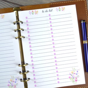 May include: A white to-do list page with a floral design at the top and bottom. The page is in a brown leather binder with a gold ring mechanism. A blue pen is lying on the right side of the page.