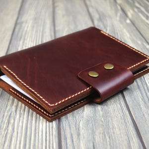 May include: A burgundy leather wallet with a flap closure secured by two brass-colored snaps. The wallet is stitched with white thread and appears to be new. The wallet is sitting on a wooden surface.